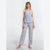 A woman modeling a two-piece pajama set consisting of a light periwinkle blue camisole top and matching wide-leg pants with a blue floral print on a white background, set against a plain white studio background.