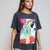 A woman modeling an oversized dark gray t-shirt with a colorful graphic print of a woman in a green dress, paired with light blue jeans, against a neutral gray background.