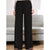 Black wide-leg pleated trousers shown on a model in a living room setting with a light gray sofa in the background.