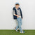 A young boy modeling a navy blue puffer vest over a navy and white striped long-sleeve shirt, paired with light blue jeans, a blue cap, and colorful sneakers, standing against a white wall with artificial green turf at the bottom.