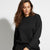 A woman wearing a black crewneck sweatshirt and matching black sweatpants, photographed against a plain light gray studio background.