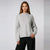 A woman modeling a light gray knit sweater with a crew neck and long sleeves, paired with black pants, against a light gray studio background.