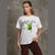A woman modeling a white graphic t-shirt featuring a green matcha drink illustration and the word 'matcha' printed on the front, paired with dark brown pants, set against a textured gray concrete wall.