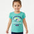 A young girl wearing a turquoise short-sleeve t-shirt with a colorful 'Flower Market' graphic on the front, paired with dark blue pants, standing against a light gray studio background.