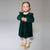 A young girl with blonde pigtails stands against a light gray studio background wearing a dark green velvet long-sleeve dress with ruffled shoulders, white knit tights, and tan lace-up shoes.