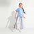 A young girl with a long braided ponytail stands in a studio wearing a light blue graphic t-shirt and blue-and-white vertically striped wide-leg pants with side slits, paired with white sneakers.
