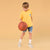 A young boy stands holding a basketball, wearing a bright yellow t-shirt with a small colorful chest graphic, denim shorts, white socks, and sneakers, set against a solid warm beige background.