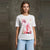 A woman modeling a white short-sleeve t-shirt with a pink and red abstract floral graphic print on the front, paired with light blue jeans, set against a textured gray concrete wall.