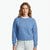 A woman wearing a heathered medium blue crewneck sweatshirt over a white undershirt and light-wash jeans, standing against a plain white studio background.