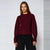 A woman modeling a dark burgundy knit crewneck sweater with black pants, standing against a light gray studio background.