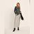 A woman models a dark gray long-sleeve knit top and a light gray maxi skirt, holding a black handbag and wearing black studded ankle boots against a plain off-white studio background.