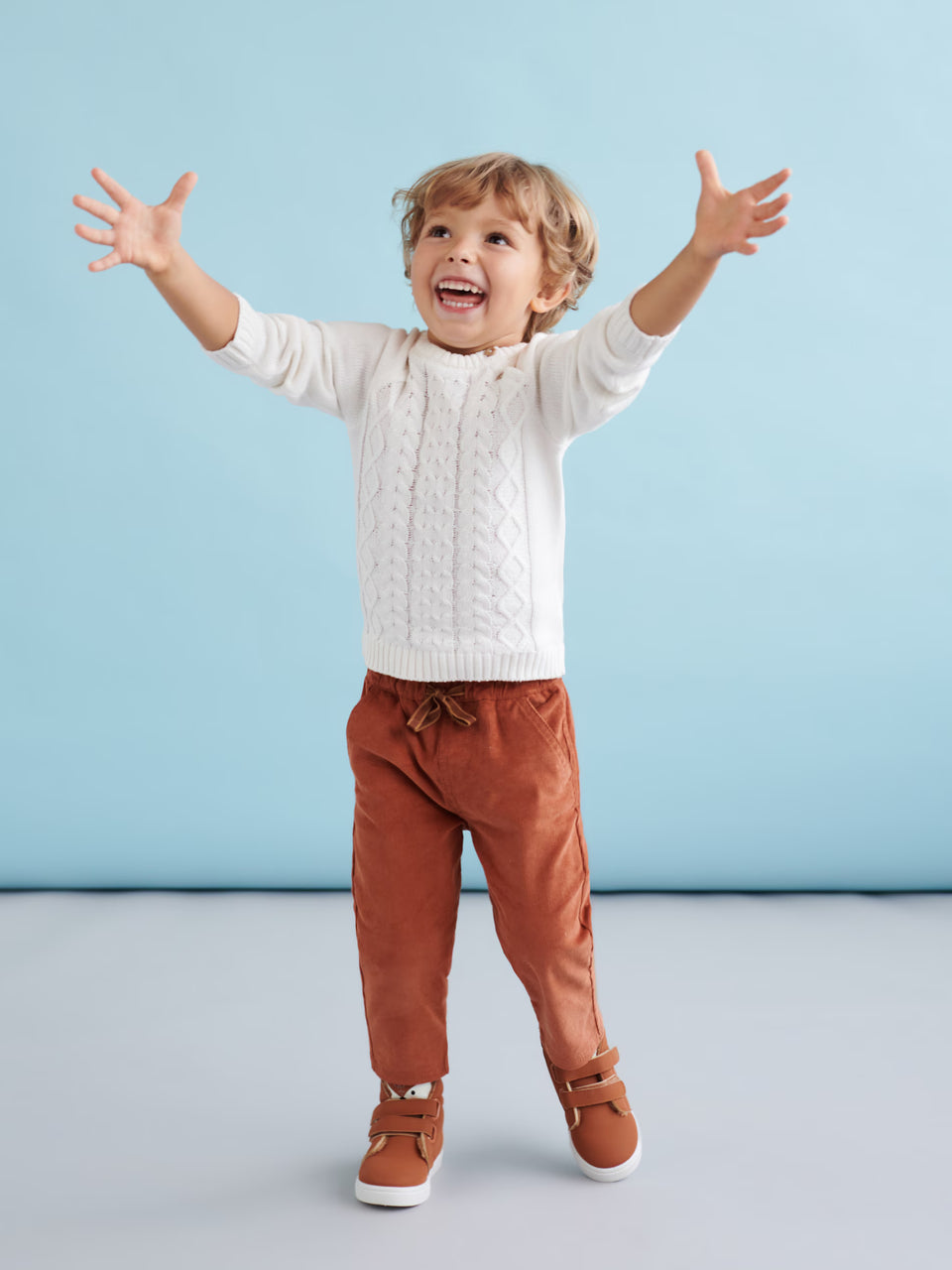 Child wearing a white sweater and brown pants against a light blue background