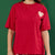 A person wearing a red short-sleeve t-shirt with a small heart patch on the chest and matching red shorts with a heart and star pattern, posed against a solid green background.