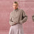 A woman wearing a chunky ribbed knit half-zip sweater in taupe and a pleated skirt, standing against a textured dusty pink background.