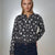 A woman modeling a dark gray knit cardigan with a white spotted pattern, paired with dark blue jeans, against a neutral gray studio background.