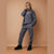 A woman modeling a dark gray lounge set consisting of a sweatshirt and jogger pants with pink embroidered details, standing against a solid tan background.