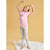 A young girl with long blonde hair is wearing a light pink short-sleeve t-shirt with a small graphic on the chest and light gray heathered leggings. She is standing barefoot on a white textured blanket against a solid warm yellow studio background.