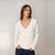 A woman wearing a white long-sleeve wrap-style sweater and dark blue jeans, standing against a light gray studio background. A small 'AI' label appears in the top-left corner.