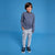 A young boy stands against a solid blue studio background wearing a navy and gray striped long-sleeve shirt, light gray jogger pants, and navy sneakers.