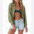 A woman modeling a sage green textured button-up shirt over a dark blue sports bra and light-wash denim shorts against a white background.