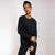 A woman modeling a black long-sleeve crewneck top and matching black pants against a light gray studio background.