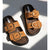 A pair of brown double-strap slide sandals with large gold-tone buckle details, displayed on a white surface with dramatic diagonal shadows.