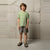 A young boy stands against a textured concrete wall wearing a light sage green short-sleeve t-shirt, gray shorts, and black slide sandals.