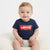 A baby sitting on a cream-colored chair wearing a navy blue Levi's short-sleeve t-shirt with the classic red and white logo on the chest, paired with a light gray diaper.
