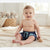 A baby sitting on a cream knit blanket in a bright room, wearing navy blue swim trunks with a mint green drawstring and a blue fish pattern. A wooden teething toy is resting near the baby's hand.