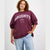A woman wearing a maroon crewneck sweatshirt with 'NAUGHTY' and a gingerbread man graphic printed in white on the front, paired with light blue jeans, against a light gray studio background.
