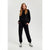 A woman modeling a black crewneck sweatshirt and matching black sweatpants with white and black sneakers, standing against a light gray studio background.