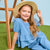 A young girl with long light brown hair sits on green artificial grass wearing a light blue textured two-piece outfit with a ruffled top and matching pants, a wide-brimmed straw hat, and posing next to a wooden ladder against a light blue backdrop.