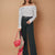 A woman models an off-the-shoulder long-sleeve striped top paired with black wide-leg pants and a black belt, holding a red handbag and a leopard-print scarf against a solid tan background.