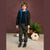 A young boy stands in a studio setting wearing a black puffer jacket with a bright blue hood and lining, a dark navy shirt, olive green pants, and navy sneakers. The background features a textured dark brown wall, a beige floor, and decorative props including a potted fern and pumpkins.