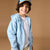 A smiling young boy wearing a light blue zip-up hoodie over a white t-shirt, paired with a blue and tan baseball cap, set against a solid warm brown studio background.