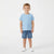 A young boy stands against a plain white studio background wearing a light blue short-sleeve crewneck t-shirt with a chest pocket, blue denim shorts, and white slip-on shoes.