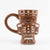 A brown ceramic mug shaped like a gingerbread person, featuring white icing-style details, a smiling face, and a handle on the left side, set against a plain white background.