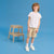 A young boy stands on a blue studio background wearing a plain white t-shirt, beige shorts with a subtle pattern, white socks, and white sneakers. A small wooden step stool is positioned to his left.