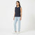 A woman modeling a sleeveless dark navy top with vertical pleats, paired with light blue jeans and white sneakers, against a plain white studio background.