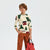 A young boy modeling a cream long-sleeve shirt with a green pine tree and red house pattern, paired with rust-brown pants, black sneakers, and holding a red backpack against a light gray studio background.