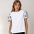 A woman modeling a white short-sleeve top with black floral lace puff sleeves, paired with black pants, against a plain light beige studio background.