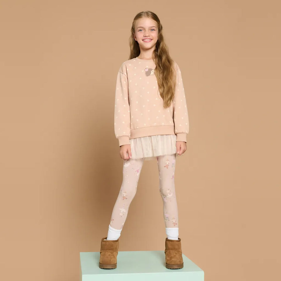 A young girl stands on a mint green pedestal wearing a light pink patterned sweatshirt, a matching pleated mini skirt, patterned leggings, white socks, and brown suede boots against a solid beige background.