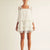 A model wearing a white tiered ruffle mini dress with cold-shoulder sleeves and light blue strappy heels, shot against a plain off-white studio background.
