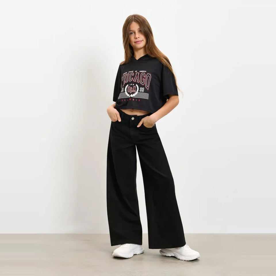 A young woman stands against a plain white wall wearing a black cropped hoodie with a vintage-style Chicago graphic, black wide-leg pants, and white sneakers.