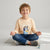 A young boy with blonde hair sits cross-legged on a light gray rug, wearing a light peach long-sleeve t-shirt with a Bluey graphic and blue jeans. A small Bluey figurine is on the floor next to him against a clean, light gray studio background.