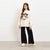 A young girl stands in a studio wearing a cream-colored sweatshirt with a colorful cartoon character graphic on the front, paired with black wide-leg pants and beige sneakers.