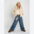 A young girl modeling a cream sherpa cropped jacket, cream knit top, wide-leg blue jeans with a plush keychain, and brown suede boots against a plain light gray studio background.