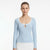 A woman modeling a light blue long-sleeve knit top with a scoop neckline, white trim, and a small keyhole cutout at the center chest, paired with a white skirt against a plain white studio background.