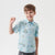 A young boy modeling a light blue short-sleeve button-up shirt with a cartoon animal print, paired with gray jeans featuring a Snoopy graphic, against a plain light gray background.