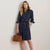 A woman models a dark blue denim shirt dress with a tie waist, paired with a tan turtleneck, brown leather handbag, and tan high heels, standing against a plain off-white wall.